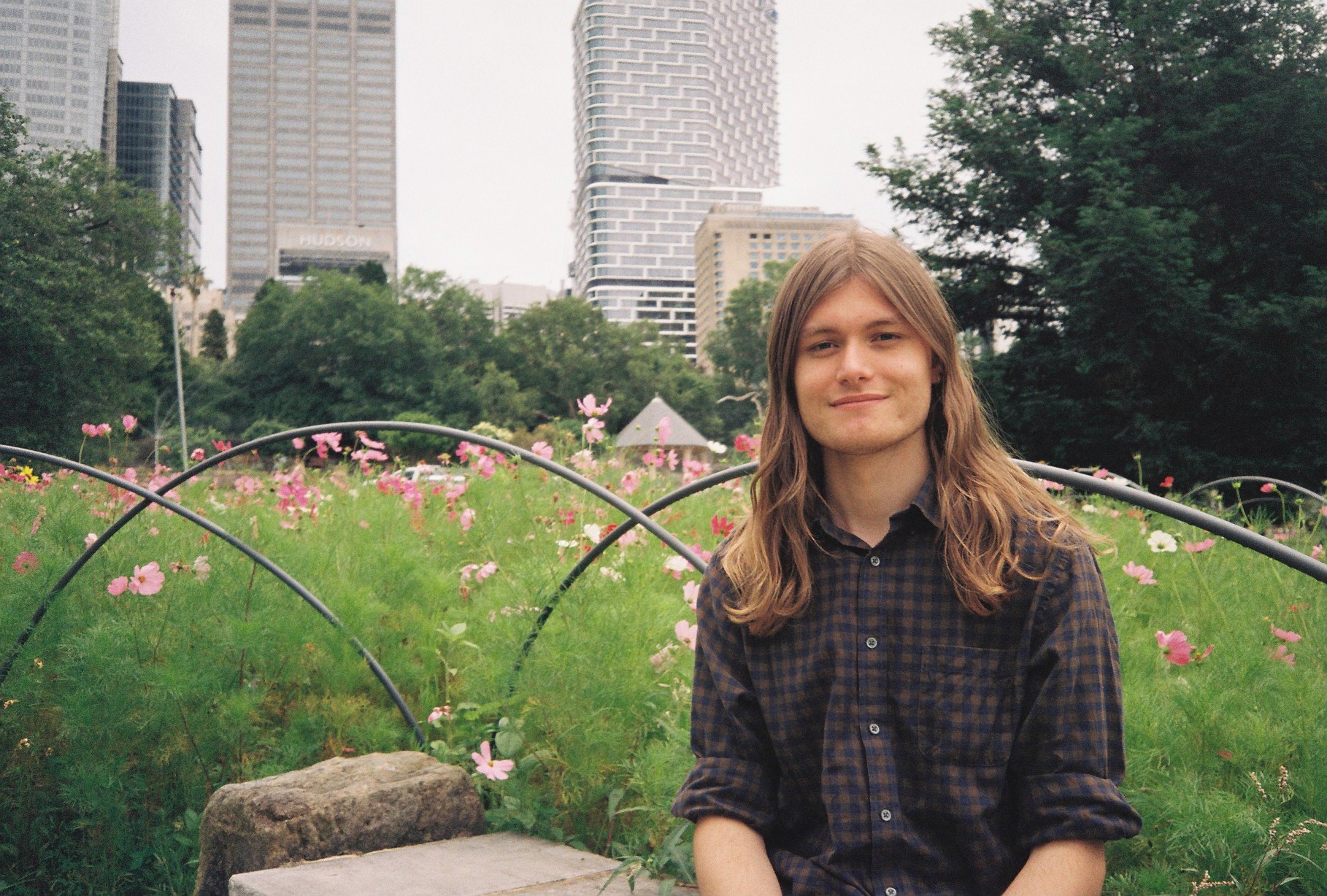 Dylan sitting on a stone in front of a fence behind which are some pink and white flowers amongst waist high bushes